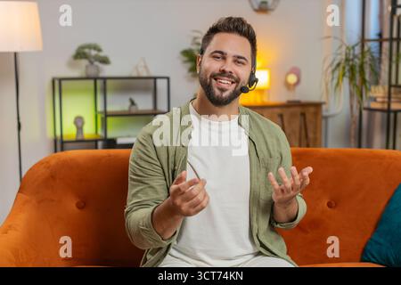 Confident young Caucasian man sitting on sofa at home wearing headset, freelance worker, call center, support service operator helpline, having talk with client or colleague, communication support. Stock Photo
