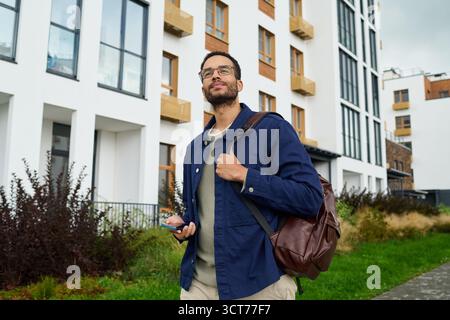 Young Black Man Walking Outdoors Holding Smartphone and Backpack Stock Photo