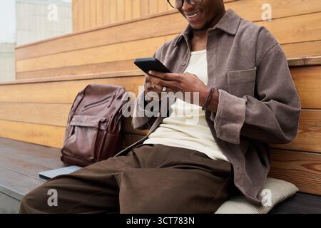 Young Black Man Sitting Outdoors Using Smartphone and Smiling Stock Photo