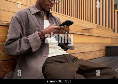 Young Black Man Sitting Outdoors Using Smartphone for Messaging Stock Photo