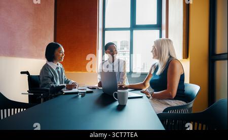 Three women, including one using a wheelchair, are conversing during a professional office meeting. Stock Photo