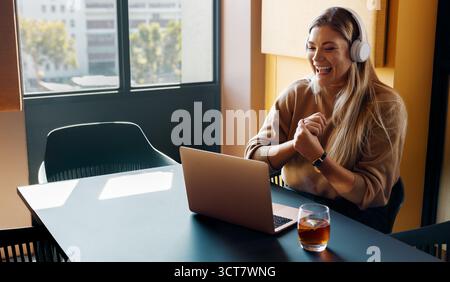 A smiling young woman wearing headphones having an engaging video call in a sunny workspace. The scene conveys professionalism, joy, and the modern ex Stock Photo
