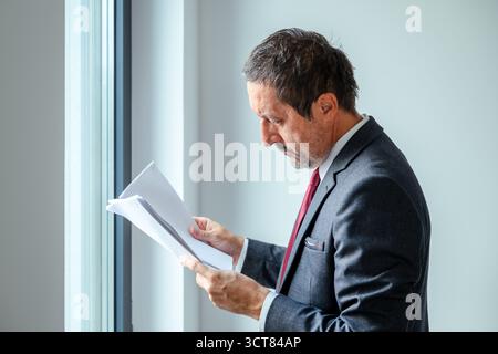 Businessman in formal suit reading documents near office window, focused professional corporate executive analyzing paper report in modern office envi Stock Photo