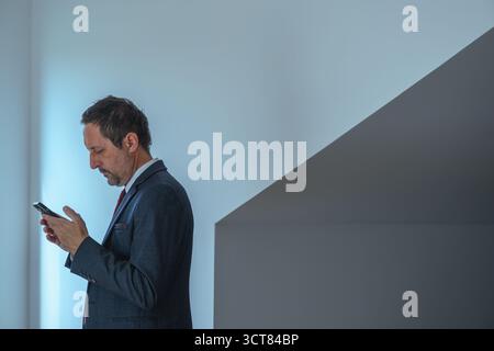 Serious businessman in suit reading text message on smartphone while standing indoors. Business leader focused on device, representing strategy, respo Stock Photo