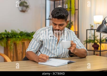 Indian man at home drinks coffee and writes goals in notebook ticks boxes and plans day with drive. Arabian guy at desk reviews list adds tasks sets priorities smiles feels focus and motivation aim Stock Photo