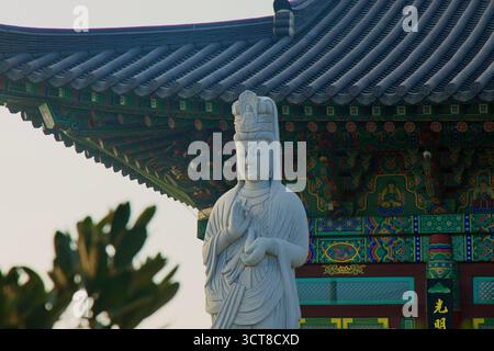 A white stone bodhisattva statue stands beside Yongmunsa Temple’s ornate eaves on Jeju’s northeast coast, with brightly painted dancheong patterns on Stock Photo