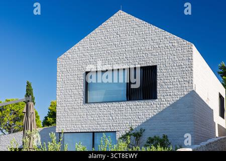 A contemporary white stone villa sits beside the coast, surrounded by green shrubs and a stone wall. Sunny day, blue sky, and tranquil seaside atmosph Stock Photo