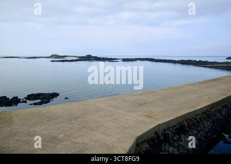 A concrete harbor wall reaches over the water toward Tokkiseom, where a sandy patch sits behind black volcanic reefs. Stock Photo
