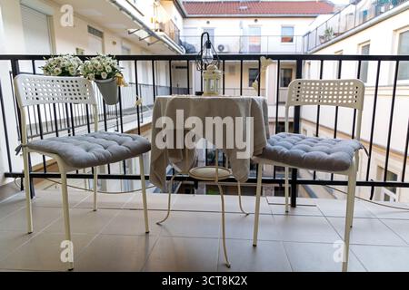 Cozy balcony setting with table and two chairs. Small balcony scene featuring a round table with beige cloth, two white chairs with gray cushions. Stock Photo
