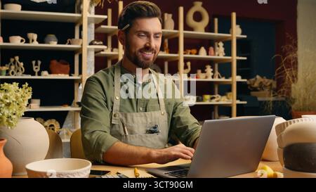 Happy joyful smiling Caucasian man small business owner craftsman businessman potter crock male working laptop typing desktop browsing computer online Stock Photo