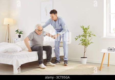 Portrait of a young friendly male nurse helping a senior patient to get off the bed at home. Stock Photo