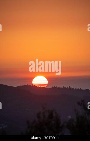 The glowing orb of the setting sun disappears behind the silhouette of a tree-lined ridge, with a hazy, warm orange and red sky above the dark hills Stock Photo