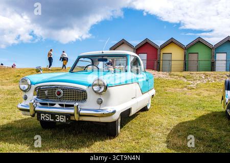 Vintage two-tone blue and white Nash Metropolitan car parked on grassy area in front of colorful beach huts under a partly cloudy sky Blyth beach Stock Photo