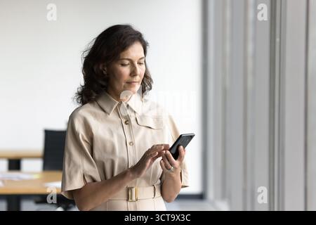 Focused mature lady entrepreneur use modern cellphone at office workplace Stock Photo