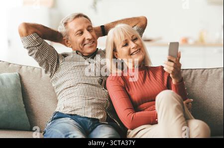 Happy Senior Spouses Using Mobile Phone Sitting On Couch Indoor Stock Photo