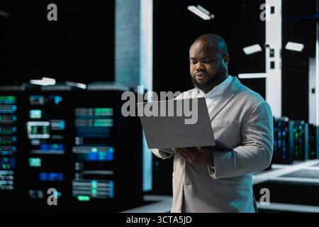Software developer in data center using laptop, reading error logs, doing checkup to prolong hardware life span. Server hub computer scientist monitoring infrastructure rigs using notebook Stock Photo