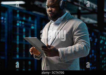 Server room IT expert listening music while fixing network issues affecting rigs performance. Data center programmer wearing audio headphones running system diagnostic tests on tablet Stock Photo