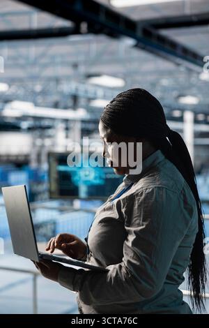 Female engineer in data center using laptop to configure hardware specifically optimized for AI tasks. Woman doing maintenance on rigs performing load balancing for artificial intelligence computing Stock Photo