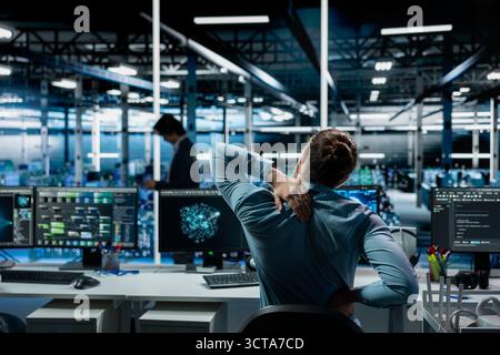 Tired data center software developer stretching to ease back tension while using AI LLM assistant. Server room IT professional massages injured back after long hours at computer coding with AI chatbot Stock Photo