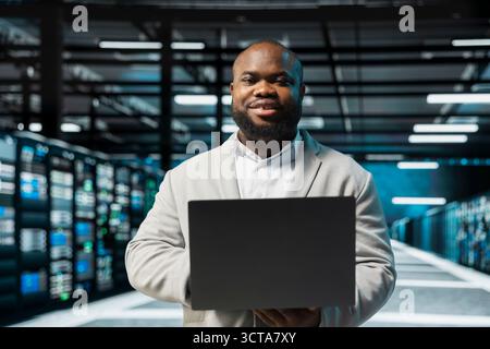 Portrait of upbeat server hub admin using laptop to fix network issues affecting storage arrays, doing backups. Jolly man in data center runs system diagnostic tests, monitoring thermal levels Stock Photo