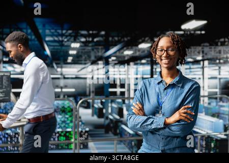 Happy IT expert in server hub reviews AI diagnostics on industrial platform, monitoring machine learning development. African american woman maintaining operational reliability, checkup. Stock Photo