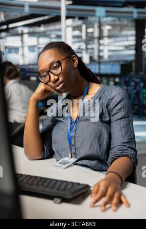 Exhausted server farm IT specialist strained after looking at computer screen for too long. Tired African american technician in data center stressed by long hardware troubleshooting session Stock Photo