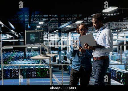 Programmers team in data center reviewing AI LLM visualizations on devices, monitoring hardware in server room. IT coworkers analyze neural network to ensure stable infrastructure operations. Stock Photo