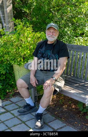 Bellevue, Washington, USA.  77 year old man in shorts with camera sitting on a park bench next to Star Jasmine (MR) Stock Photo