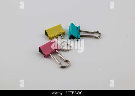 Three vibrant binder clips in pink, yellow, and teal are laid out on a neutral surface, showcasing their utility for organizing papers and documents. Stock Photo