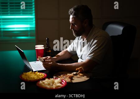 Businessman eats a late-night snack while working on laptop. A tired employee using laptop on late-night overtime. Latin Business man overworked into Stock Photo