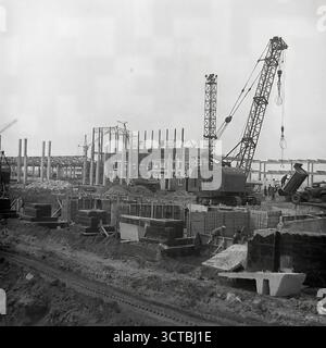 A panoramic view of a massive industrial construction site in 1960s Sloviansk, Ukrainian SSR. This archival photo shows the scale of Soviet ambition, with the vast concrete framework of a new factory rising from the ground. Heavy cranes, including a prominent MKG-25 model, dominate the landscape as they lift prefabricated concrete sections. The scene captures the essence of the era's focus on heavy industrialization in the Donbas region. This powerful image symbolizes the immense activity, progress, and industrial might of the time Stock Photo