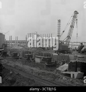 A panoramic view of a massive industrial construction site in 1960s Sloviansk, Ukrainian SSR. This archival photo shows the scale of Soviet ambition, with the vast concrete framework of a new factory rising from the ground. Heavy cranes, including a prominent MKG-25 model, dominate the landscape as they lift prefabricated concrete sections. The scene captures the essence of the era's focus on heavy industrialization in the Donbas region. This powerful image symbolizes the immense activity, progress, and industrial might of the time Stock Photo