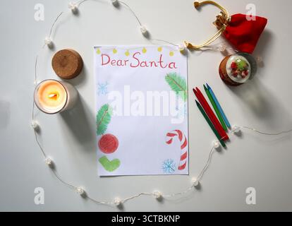 Flat lay of a Dear Santa letter with Christmas decorations, burning candle, red pouch, garland, crayons and snow globe on white table. Letter template Stock Photo
