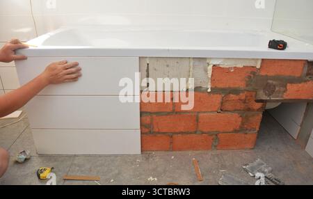 Facing the bathtub with white ceramic tiles. Builder installing ceramic tiles on bathtub wall. Stock Photo