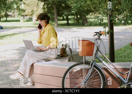 Middle Aged Caucasian Woman Working on Laptop and Talking on Smartphone Outdoors Stock Photo