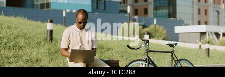 Young Adult Black Man Sitting Outdoors Using Laptop near Bicycle on Grass Stock Photo