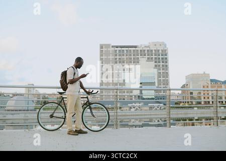 Young Adult Black Man Standing Outdoors Using Smartphone Beside Bicycle Stock Photo
