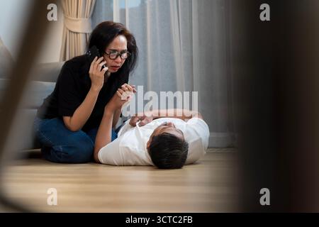 A worried middle-aged woman is calling emergency services on her smartphone while kneeling beside an unconscious man lying on the floor at home. This Stock Photo