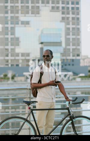 Young Adult Black Man Standing Outdoors Holding Smartphone Beside Bicycle Stock Photo