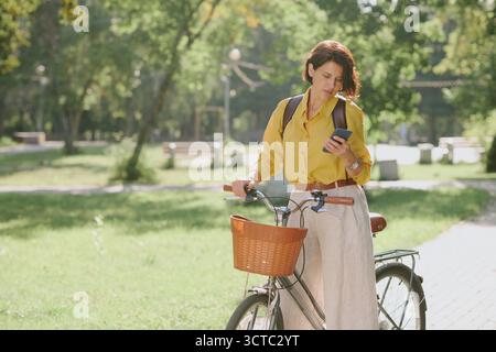 Caucasian Middle Aged Woman Walking Bicycle While Checking Smartphone Outdoors Stock Photo