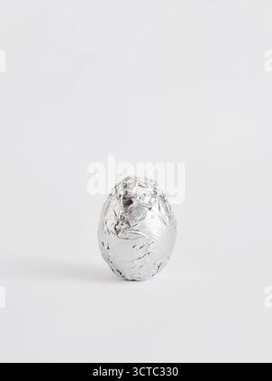 Easter egg wrapped in shiny silver aluminum foil, minimalist food concept on a plain white background, still life image Stock Photo