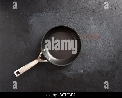 Empty frying pan featuring a non-stick surface and stainless steel handle, positioned on a textured dark gray cooking surface Stock Photo