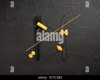 Pasta assortment in varied uncooked shapes with a single spaghetti strand threaded through black fork and knife on black background Stock Photo