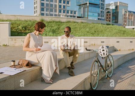 Young Caucasian Woman and Young Black Man Discussing Blueprints Outdoors Stock Photo