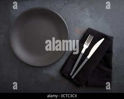 Empty black plate and cutlery on a black napkin, preparing minimalist table setting on dark stone background Stock Photo