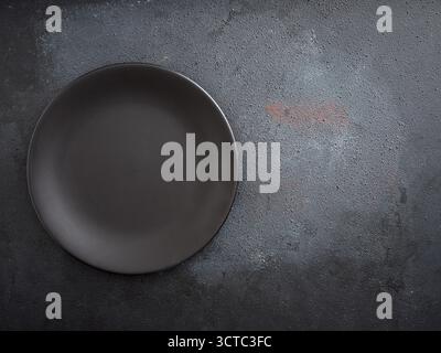 Empty round black ceramic plate with a matte finish resting on a dark, textured gray stone or concrete surface Stock Photo