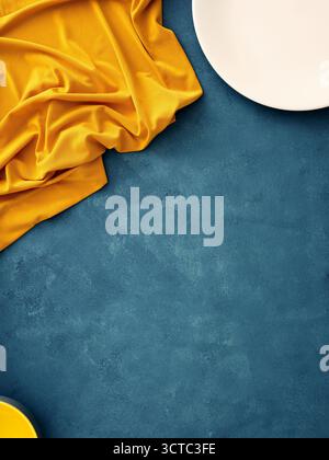 Table setting featuring a white plate, fork, knife, and yellow napkin on a blue background. Minimalist flat lay for food mockup setting with copy spac Stock Photo