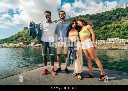 Happy group of four young diverse friends standing together and smiling at the camera for a portrait on a seaside pier during a summer holiday in Ital Stock Photo