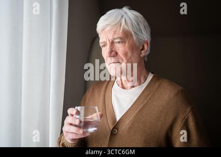 Sad sorrowful old man drinking water from glass looking out window. Loneliness and aging disease Stock Photo