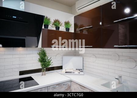 A trendy kitchen with white countertops, an induction hob, and spotlights. A contemporary corner kitchen with white wood cabinets and glossy brown cab Stock Photo